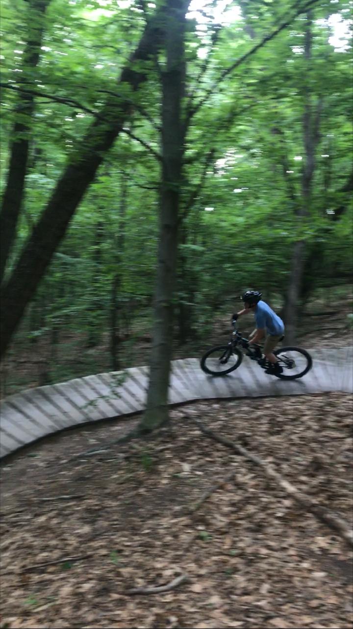 A person riding a mountain bike on a curved wooden trail in a lush green forest, surrounded by trees and leaf-covered ground. The image captures the motion of biking, with the rider leaning into the turn. Merrell Trail mountain bike trail.