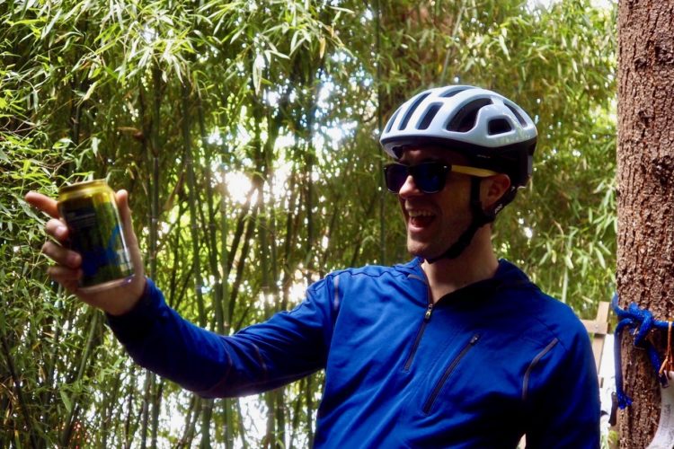 A person wearing a white bicycle helmet and dark sunglasses is smiling and holding a can of beverage, standing in a lush green area with bamboo in the background. They are dressed in a blue zip-up jacket.