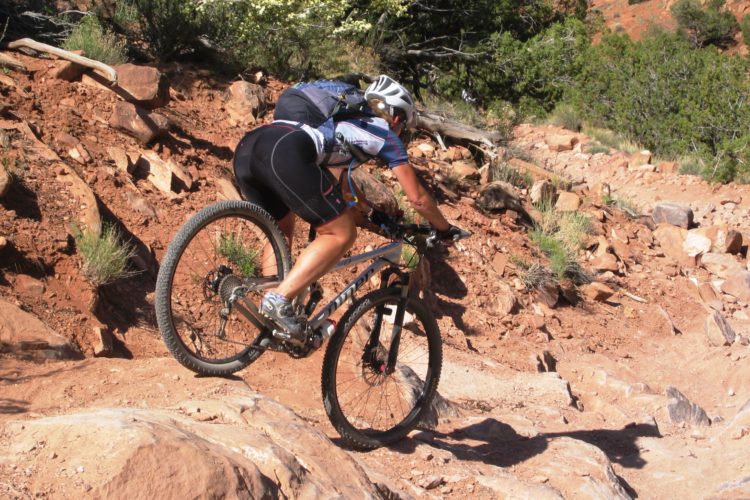 A mountain biker navigating a rocky trail, wearing a helmet and cycling gear, with the bike's front wheel lifted slightly off the ground. The background features a rugged landscape with red earth and sparse vegetation.