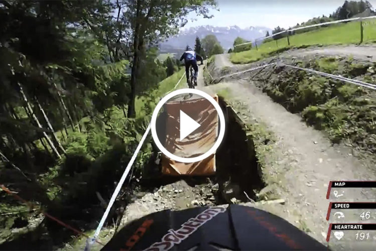 A view from a mountain bike's perspective as it approaches a jump on a dirt trail, surrounded by trees and greenery. The screen displays real-time metrics such as speed (46 km/h) and heart rate (191 bpm). A video play icon is centered in the image, indicating video content.