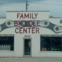 A storefront of a bicycle shop called "Family Bicycle Center," featuring bold red letters on a white background. The building has large windows displaying bicycles and accessories, with a recognizable design element resembling bicycle gears on either side of the name. The address "3410" is visible beneath the store