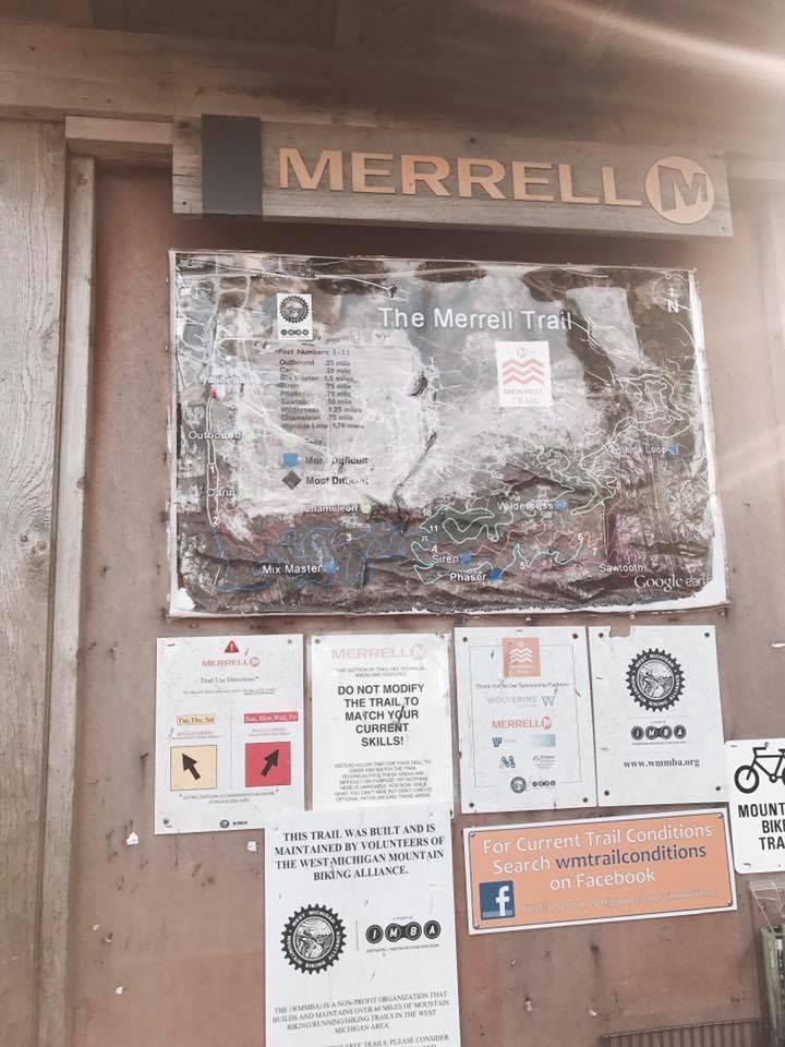 A wooden signboard displaying a map of the Merrell Trail, along with various informational posters about trail guidelines and maintenance. The map highlights different trail sections, difficulty levels, and major landmarks. Additional signs include tips for trail users, instructions to not modify the trail, and contact information for trail conditions on social media. The overall setting suggests a trailhead for hiking or biking activities. Merrell Trail mountain bike trail.