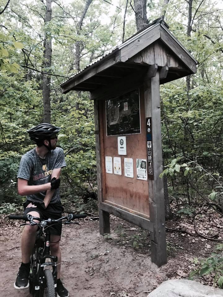 A person wearing a helmet and cycling gear sits on a mountain bike, looking at a wooden information board in a forested area. The board displays maps and various signs related to trails and outdoor activities. Lush green trees surround the scene, indicating a natural setting suitable for biking and hiking. Merrell Trail mountain bike trail.