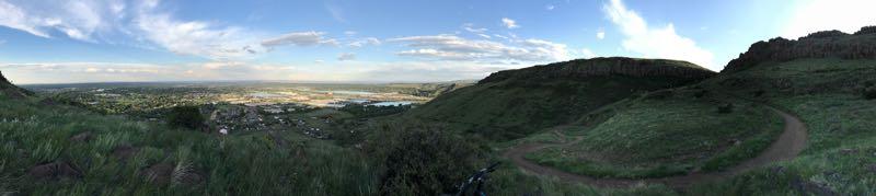 Panoramic view of a lush green landscape with rolling hills, a winding path, and a body of water visible in the distance. The sky is partly cloudy with blue and white hues, capturing the beauty of nature during daylight. North Table Mountain mountain bike trail.