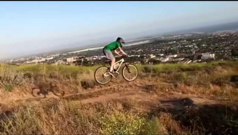 A person in a green shirt and gray shorts rides a mountain bike off a small dirt jump, with a view of a city and the ocean in the background. The scene captures a sunny day with clear skies and lush greenery surrounding the trail. Sanjon Barranca mountain bike trail.