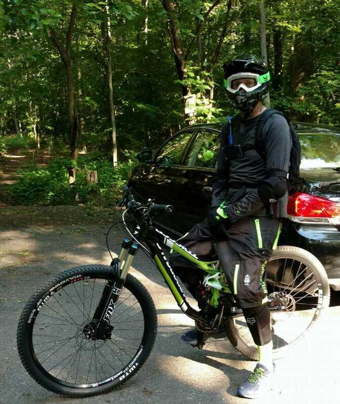 A person dressed in a full-face helmet, goggles, and protective gear stands next to a mountain bike on a dirt path surrounded by trees. They are wearing a black outfit with green accents and appear ready for a biking adventure. A black car is parked in the background. Rancocas State Park - Westampton mountain bike trail.