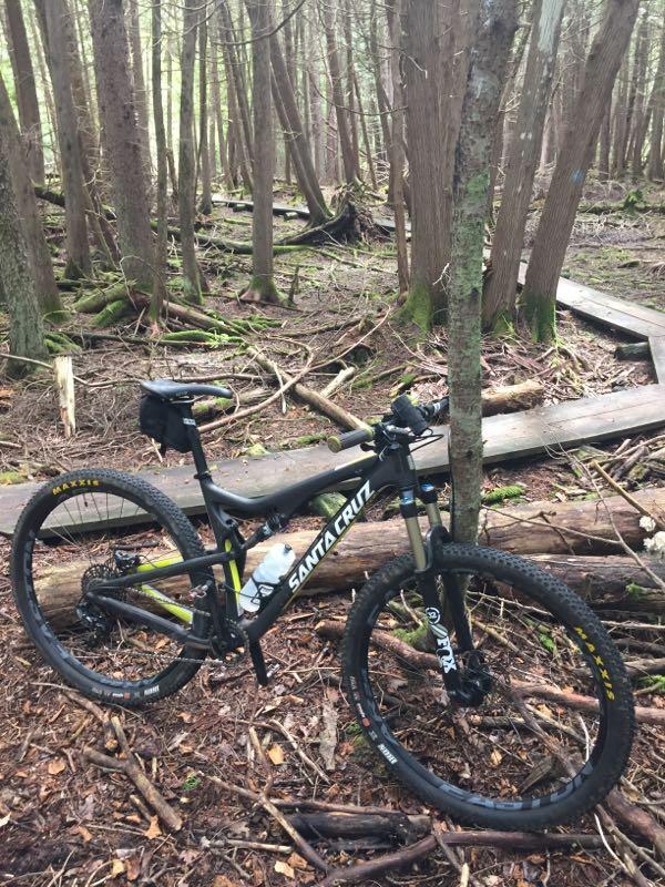 A black mountain bike leaning against a tree in a dense forest. The ground is covered with fallen leaves and branches, and there is a wooden pathway in the background. The bike features a recognizable brand name, Santa Cruz, on the frame. Shingle Mill Pathway mountain bike trail.