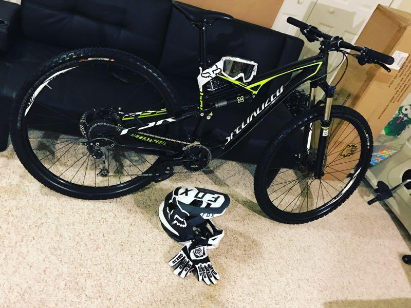 Mountain bike leaning against a couch, accompanied by a helmet and gloves. The bike features a black and green design, with prominent tires and gearing components. The setting includes a carpeted floor and a dark couch in the background. Brighton Rec Area mountain bike trail.