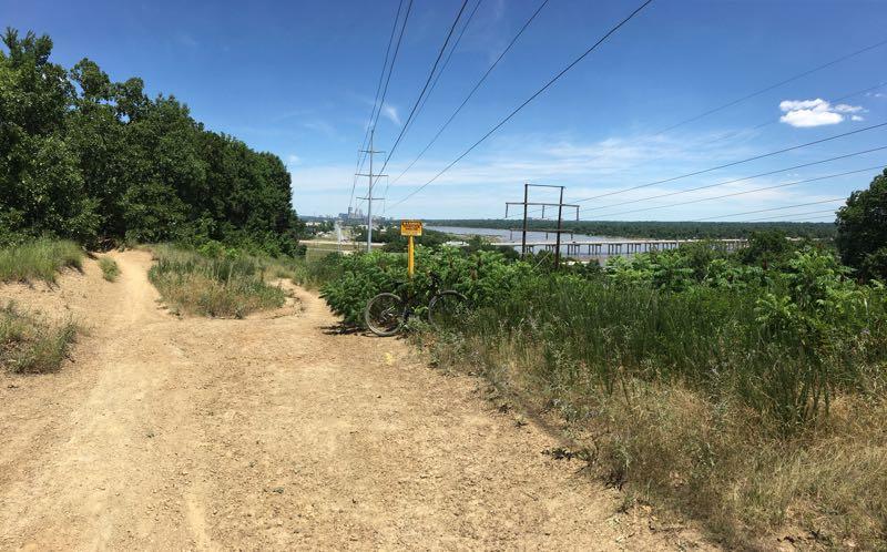 A dirt path leading through tall grass and bushes, with power lines overhead. A bike is parked at a sign indicating a nearby trail, and the landscape includes a river and distant structures under a clear blue sky. Turkey Mountain mountain bike trail.