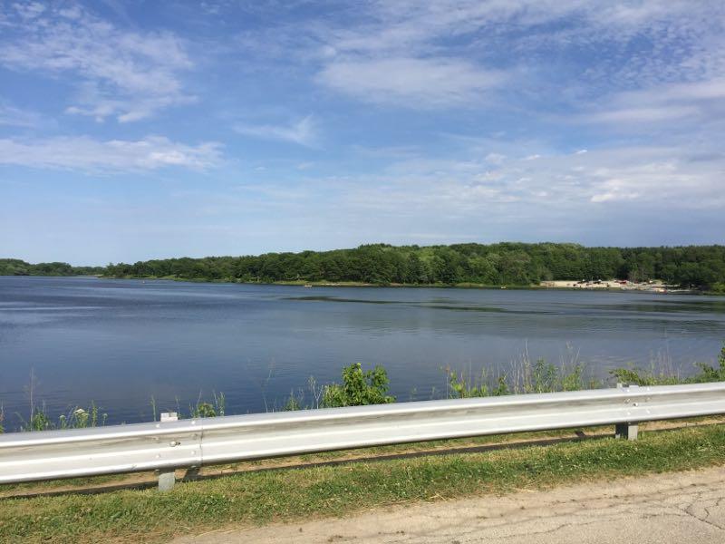 A serene view of a calm lake surrounded by lush greenery under a partly cloudy sky, with a white guardrail in the foreground and hints of recreational areas visible along the shore. Rock Cut State Park mountain bike trail.