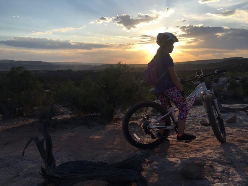 A young person in cycling gear stands next to a mountain bike, silhouetted against a sunset. The scene features expansive desert landscapes with distant mountains and scattered greenery, creating a serene outdoor atmosphere. Dead Horse Point State Park mountain bike trail.