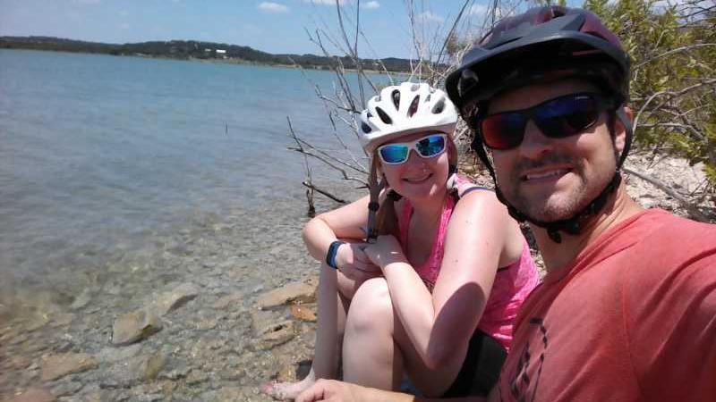 A smiling couple sits by the water's edge on a sunny day, wearing vibrant athletic clothing and helmets. The woman has a white helmet and sunglasses, while the man is wearing a black helmet and sunglasses. They are surrounded by rocks and greenery, with a calm lake in the background under a clear blue sky. Madrone Trail mountain bike trail.