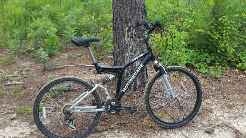 A black and silver Motobecane mountain bike is leaning against a tree in a natural, wooded setting. The bike features thick tires and a sturdy frame, surrounded by green plants and pine needles on the ground. Children's Home / Pig Trail mountain bike trail.