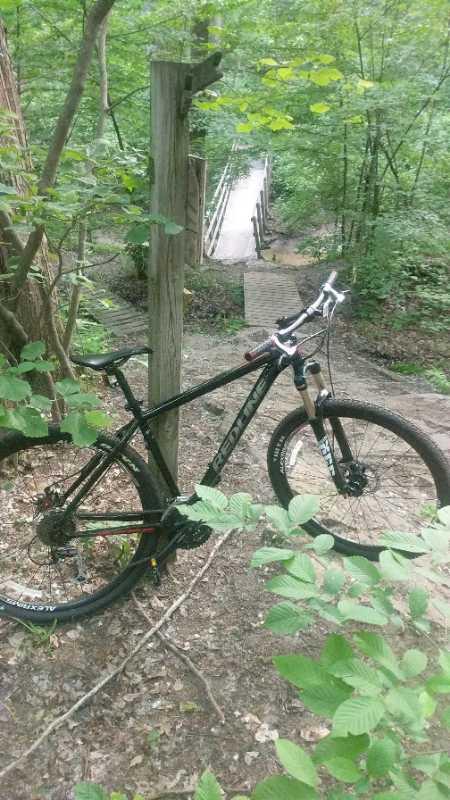 A black mountain bike leaning against a wooden post, surrounded by lush green foliage, with a wooden bridge visible in the background, leading into a dense forest. Midland City Forest mountain bike trail.