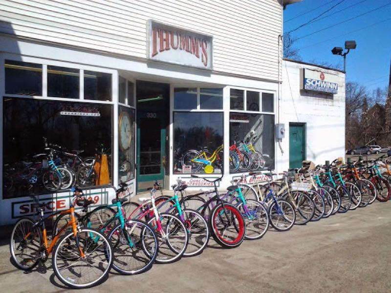 A storefront of a bicycle shop named "Thom's" with a display of various colorful bicycles lined up outside. The shop features large windows showcasing additional bikes inside, and a sign for "Schwinn" is visible on the adjacent building. The image captures a sunny day with clear skies.