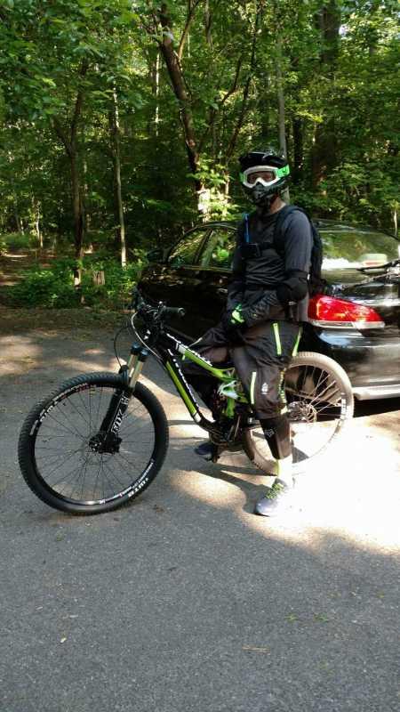 A person wearing a black helmet, goggles, and protective gear stands next to a mountain bike on a gravel path in a wooded area. A parked car is visible in the background. The scene is surrounded by green trees. Rancocas State Park - Westampton mountain bike trail.