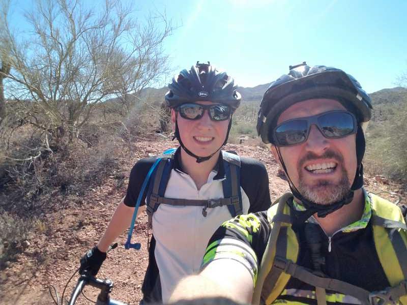 A man and a woman pose for a selfie while mountain biking in a sunny desert landscape. Both are wearing helmets and sunglasses, and the woman is dressed in a white and black cycling jersey, while the man sports a colorful striped shirt. In the background, there are sparse shrubs and rocky terrain, with mountains visible under a clear blue sky. Hawes Loop mountain bike trail.
