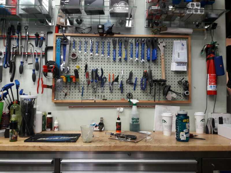 Alt text: A workshop bench with a pegboard displaying various tools such as wrenches, pliers, and screwdrivers. The work surface is cluttered with bottles, tools, and a cup, while additional tools and equipment are visible on shelves above.