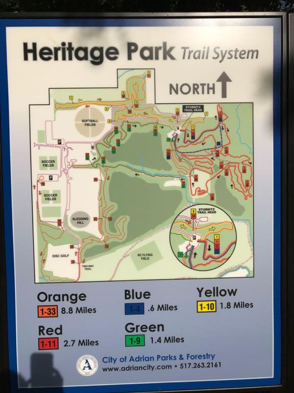 Map of the Heritage Park Trail System, displaying various trails marked by color (Orange, Blue, Yellow, Red, Green) along with their respective mileages. Includes landmarks such as soccer fields, softball fields, a sledding hill, a disc golf area, and the Stunitz Trail Head. The map orientation indicates north at the top. Heritage Park mountain bike trail.