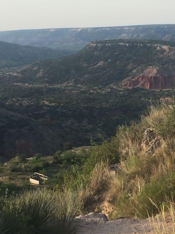 A panoramic view of a canyon landscape featuring rolling hills and rocky cliffs, with green vegetation in the foreground and hints of red rock formations in the distance under a clear sky. A wooden observation platform can be seen nestled in the valley below. Palo Duro Canyon mountain bike trail.