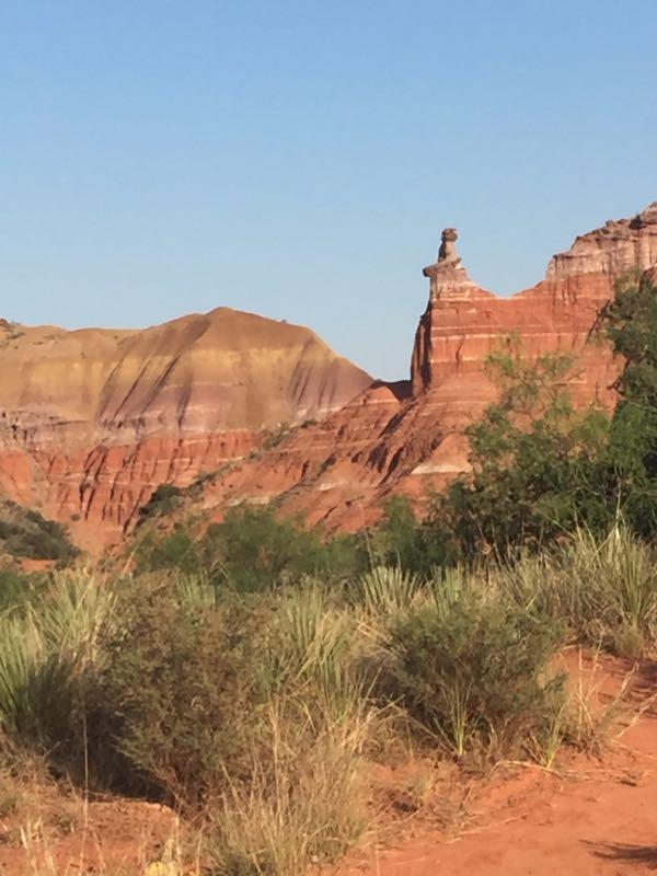 A scenic view of a rugged landscape featuring layered red rock formations and vegetation in the foreground. A prominent rock formation resembling a figure stands on the edge of one of the cliffs under a clear blue sky. Palo Duro Canyon mountain bike trail.
