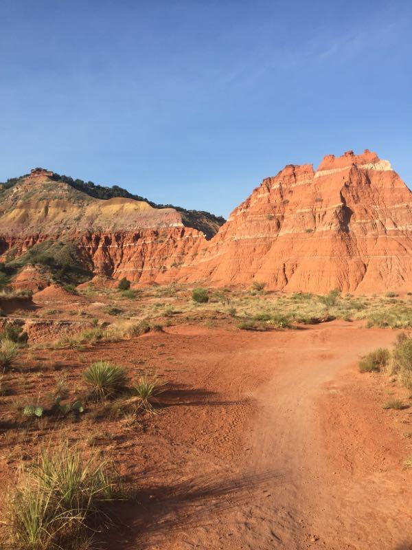 A scenic view of a rugged landscape featuring striking red rock formations and steep hills, with a clear blue sky above. A winding dirt path leads through the arid terrain, surrounded by sparse vegetation and grasses typical of a desert environment. Palo Duro Canyon mountain bike trail.