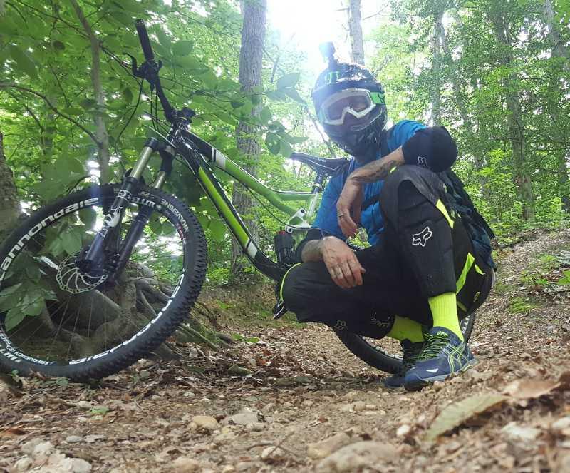 A person in cycling gear kneels beside a mountain bike in a forested area. The individual is wearing a helmet with a camera attached and protective clothing, including a blue shirt and black pants with yellow accents. The background features trees and foliage, creating a lush outdoor setting. Camden County College mountain bike trail.