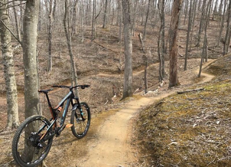 A mountain bike resting on a dirt trail in a wooded area. The scene features bare trees in early spring, with winding paths leading through the forest. The ground is covered in a mix of dirt and sparse foliage, suggesting a natural outdoor setting. Fountainhead Regional Park mountain bike trail.