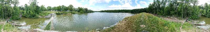 A panoramic view of a serene lake surrounded by lush greenery and trees under a partly cloudy sky. The calm water reflects the surrounding landscape, creating a peaceful, natural atmosphere. A grassy bank leads to the water's edge, enhancing the tranquil setting. Van  Buren mountain bike trail.