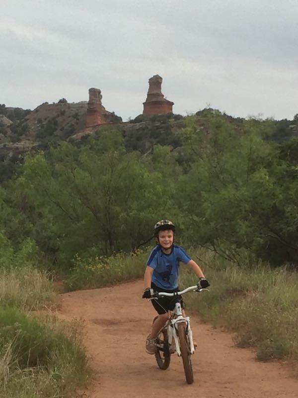 A young boy wearing a helmet rides a white bicycle on a dirt path surrounded by green vegetation, with rock formations visible in the background under a cloudy sky. Palo Duro Canyon mountain bike trail.