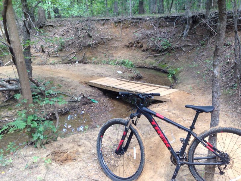A mountain bike is positioned on the sandy bank beside a small creek, with a wooden bridge crossing over the water. Surrounding the scene are trees and underbrush, indicating a wooded area. The path leading to the bridge is uneven and natural, suggesting a trail in a forested setting. Northshore Trail mountain bike trail.