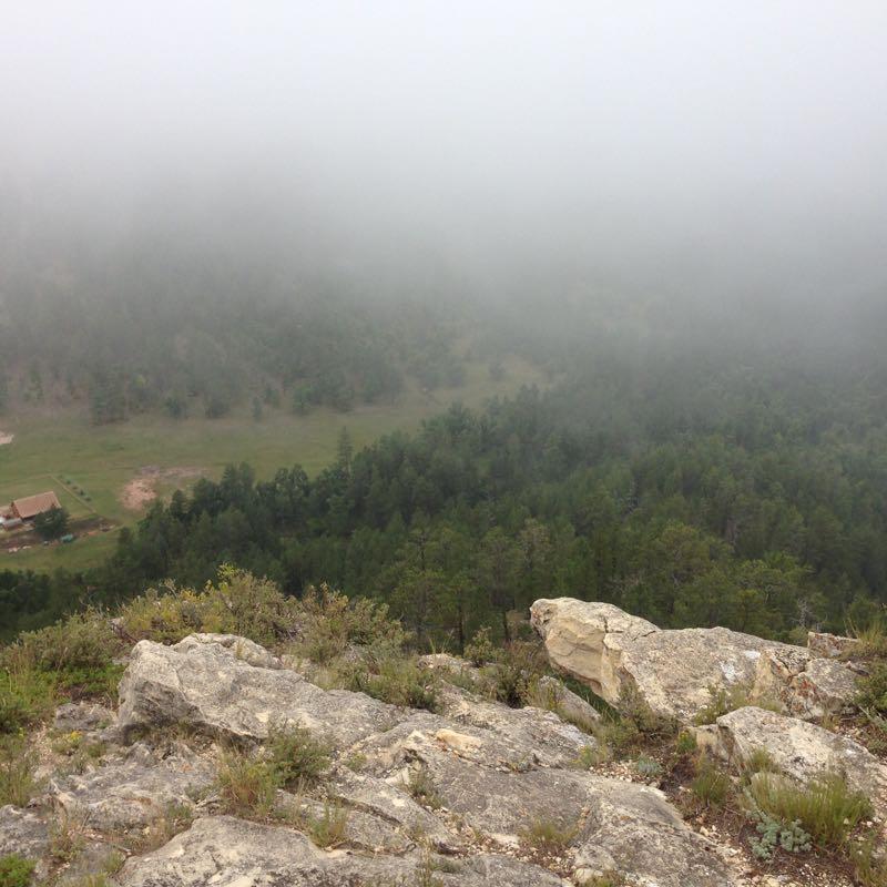Foggy landscape view from a rocky cliff, overlooking a forested area with a small building partially visible in the valley below. The scene conveys a sense of tranquility and isolation, with dense fog obscuring much of the surrounding greenery. Victoria's Secret mountain bike trail.