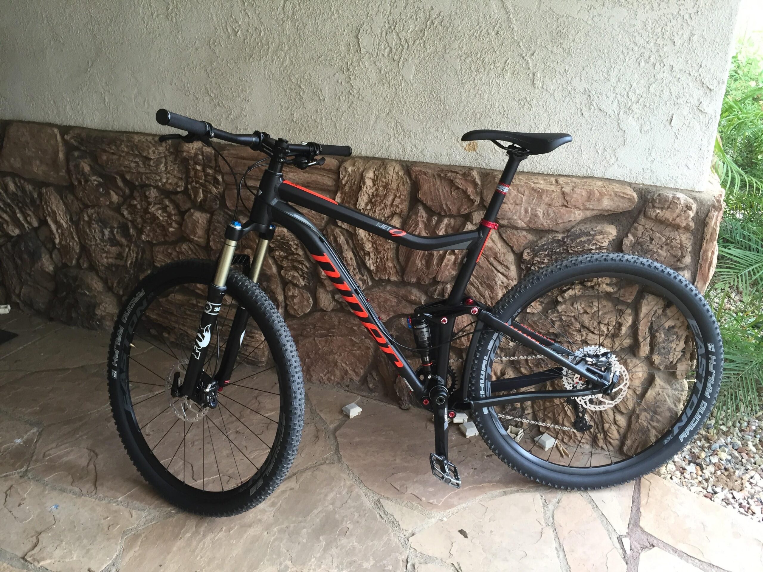 Niner Jet 9: A black and red mountain bike leaning against a stone wall, featuring thick tires, a front suspension fork, and a sleek frame design. The ground is made of stone tiles, and there are some plants visible in the background.