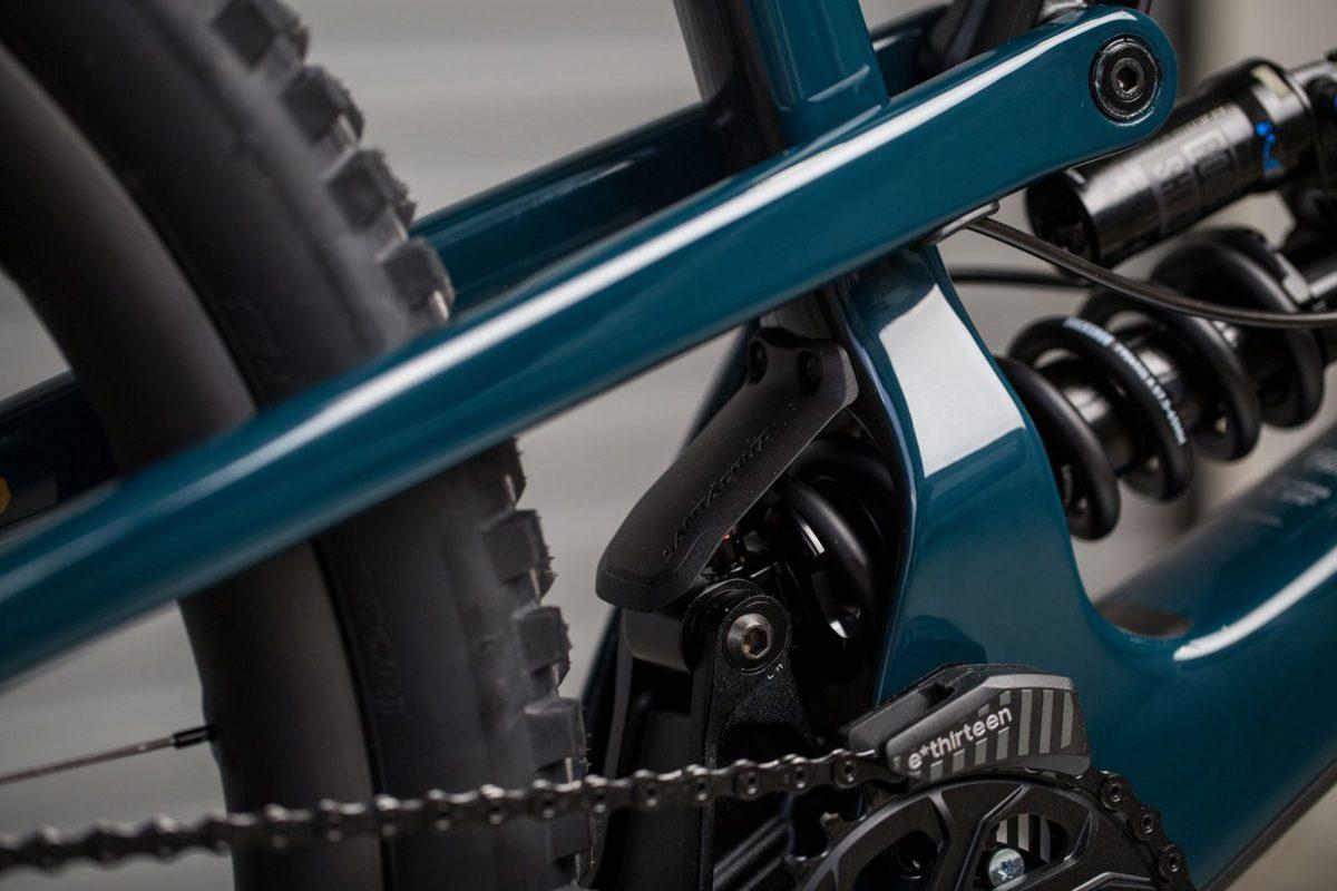 Santa Cruz Nomad: Close-up view of a mountain bike's rear suspension system, showcasing the teal frame, coil spring, and derailleur. The image highlights the intricate mechanics, including the chain and tire tread.