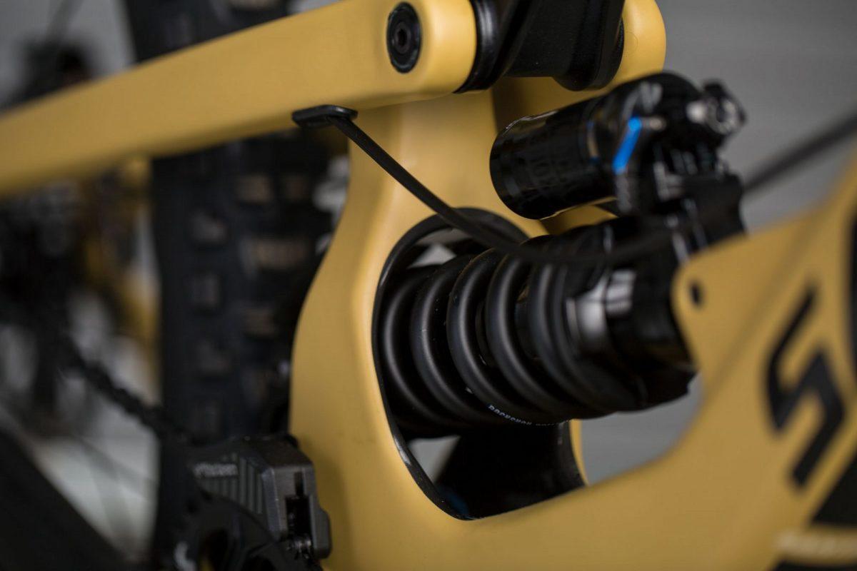 Santa Cruz Nomad: Close-up of a bicycle's rear suspension system featuring a black spring and shock absorber mounted on a yellow frame, with visible components like the chain and gears in the background.