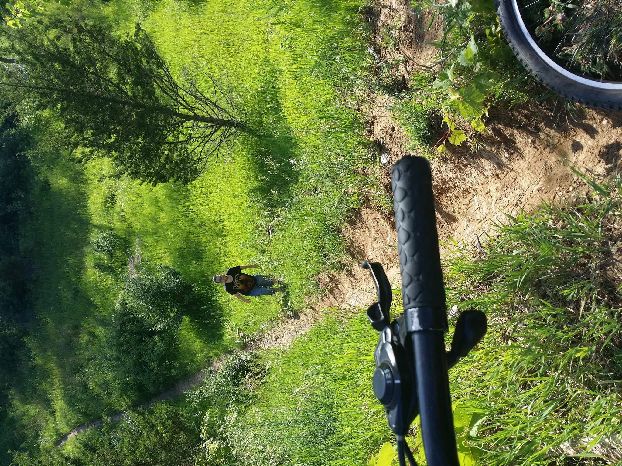 A perspective view of a mountain biking trail with a bicycle handlebar in the foreground. A person is walking along a dirt path surrounded by tall green grass and trees in a sunny outdoor setting. Raceway Woods mountain bike trail.