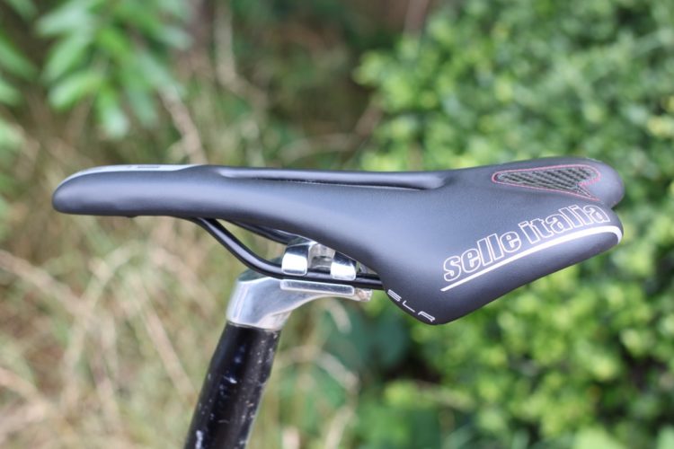 Close-up of a black Selle Italia SLR bike saddle, attached to a silver saddle rail, with green foliage in the background.