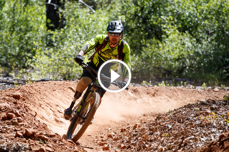 A mountain biker in a bright green jersey navigates a dirt trail, kicking up dust as he leans into a turn among lush green trees.