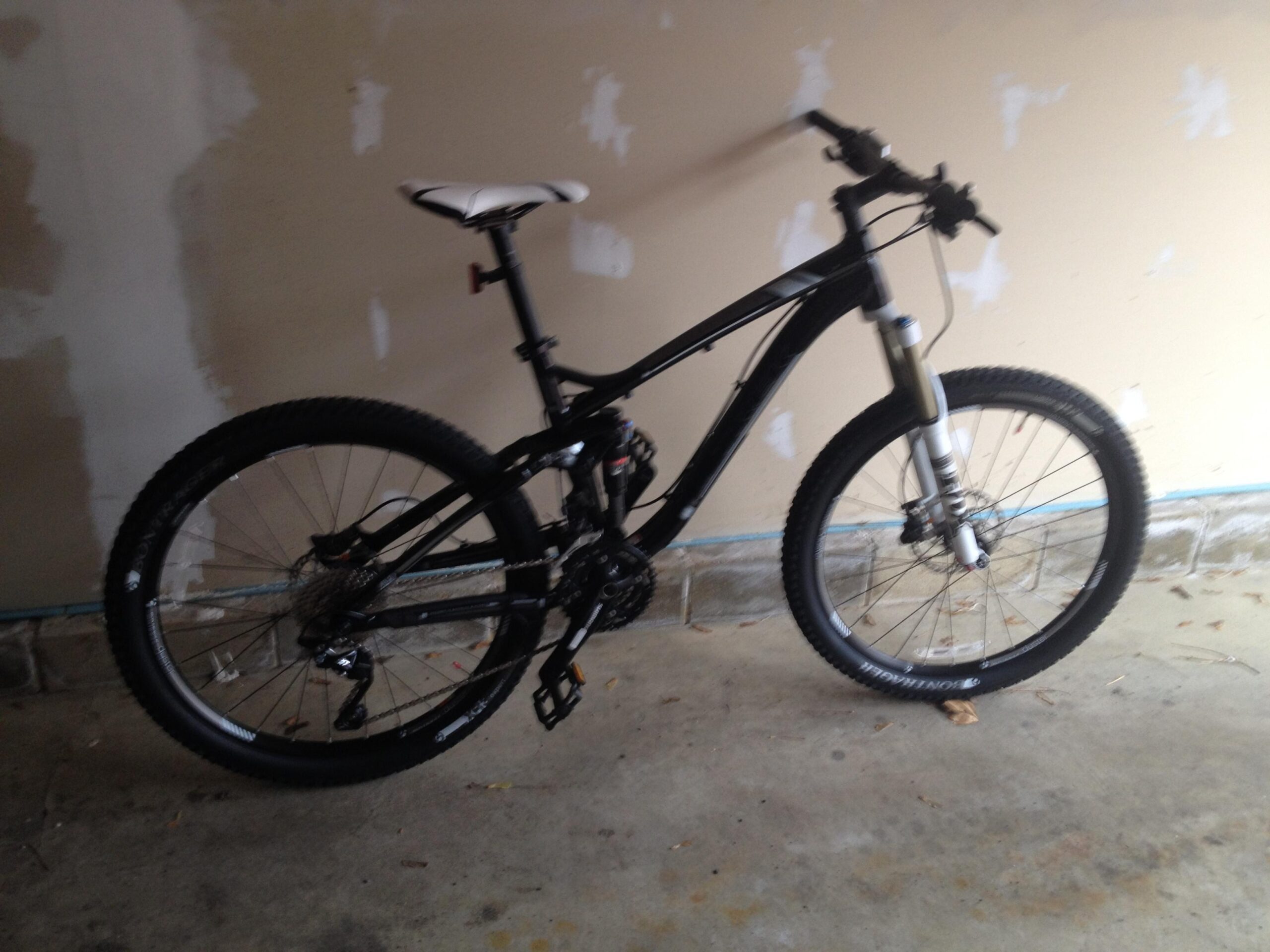 Trek Fuel EX 8: A black mountain bike with a white seat, positioned in a garage with a plain wall in the background. The bike features thick tires, a suspension fork, and a gear system.