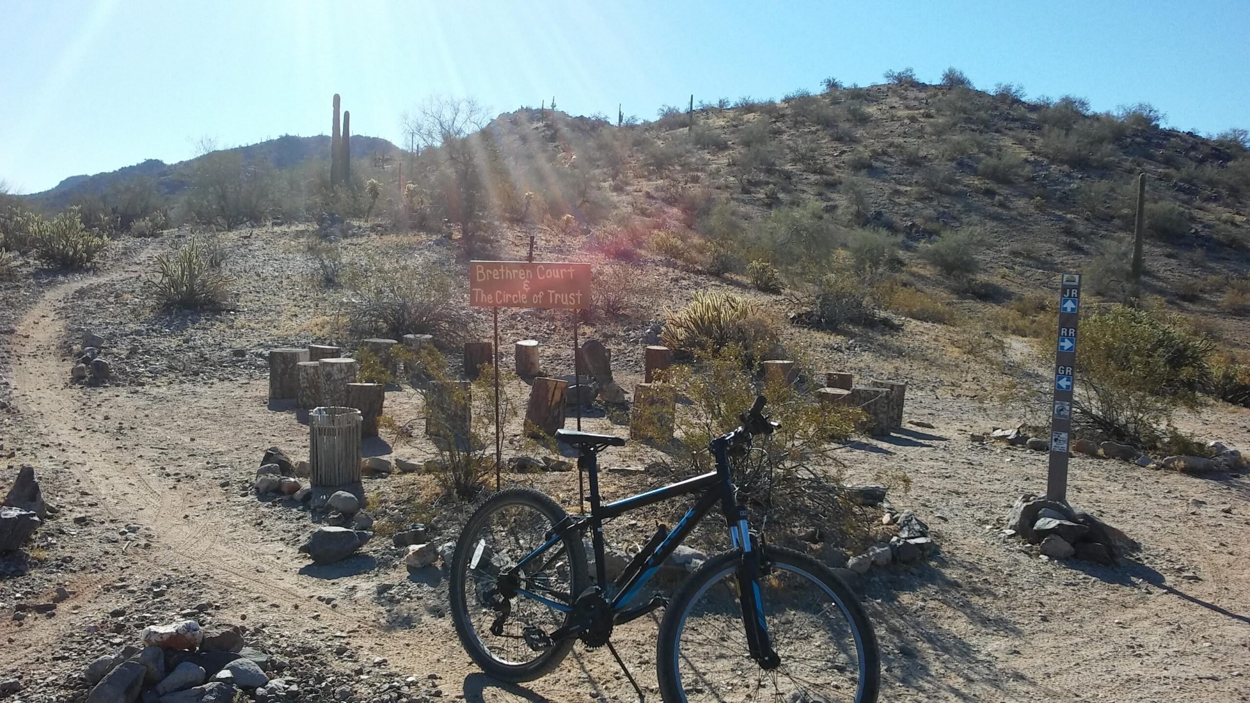 Trek 820: A black mountain bike is positioned on a dirt path in the desert, with a sign in the background reading "Brethren Court & The Circle of Trust." The landscape features rocky terrain, desert vegetation, and hills under a clear blue sky. Trail markers indicating different routes are visible nearby.
