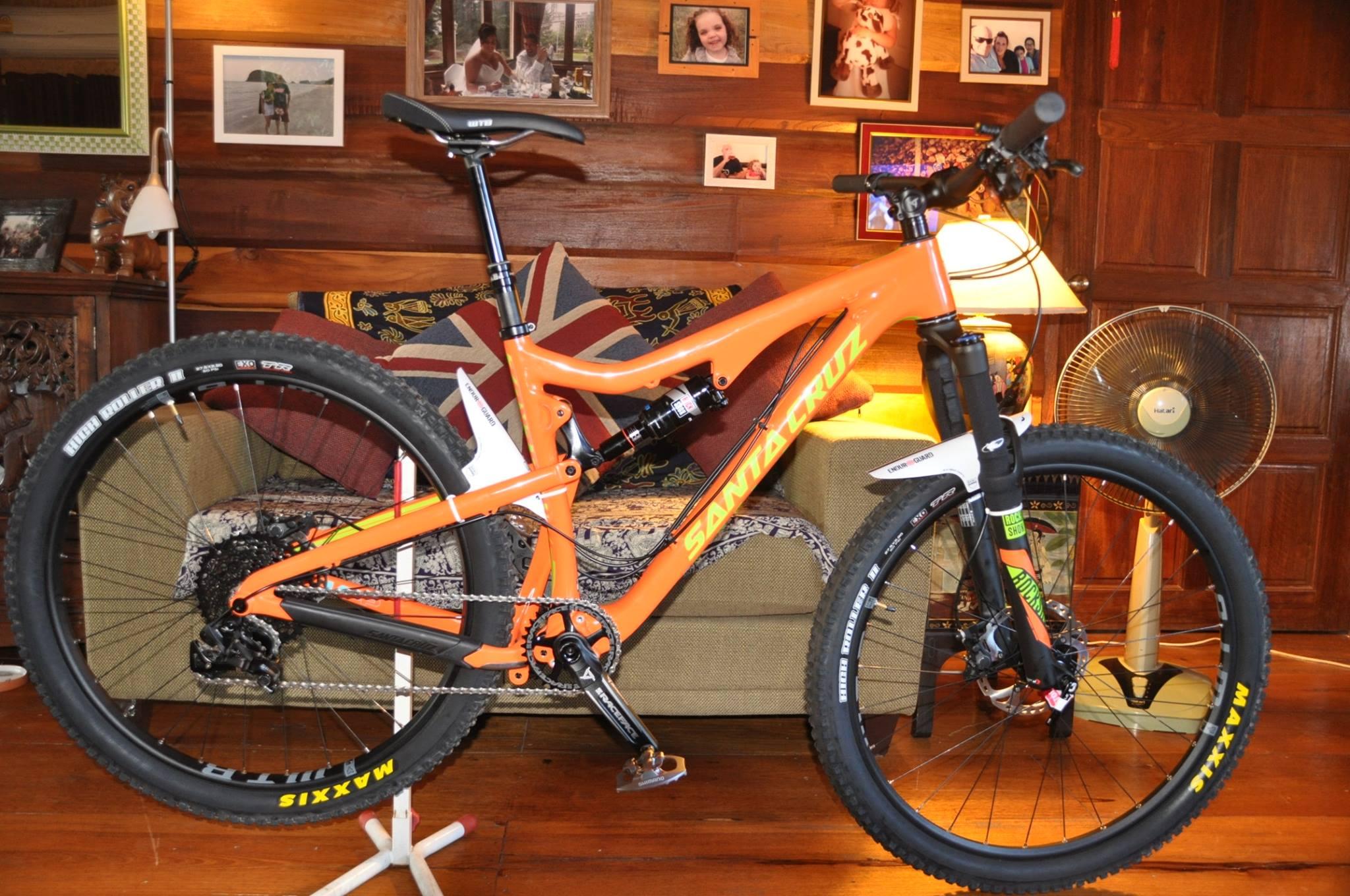 Santa Cruz 5010: A bright orange mountain bike positioned indoors, featuring a sleek frame, wide tires, and visible components from the manufacturer Santa Cruz. In the background, there is a cozy living room setting with a green couch, decorative cushions, and various framed photos on the wall. A standing fan is also visible, contributing to the relaxed home environment.