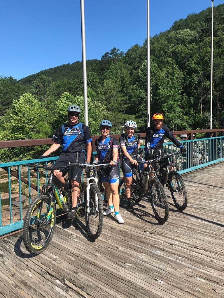 Four mountain bikers pose for a photo on a wooden bridge, surrounded by lush green trees. They are wearing matching blue and black cycling jerseys and helmets. Each person is standing beside their mountain bike, showcasing a sunny day and a scenic outdoor environment. Tanasi Trail System mountain bike trail.