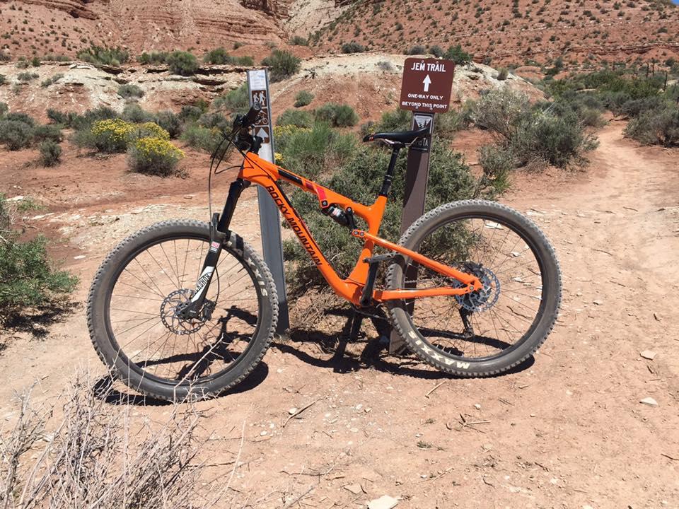 Rocky Mountain Pipeline: A mountain bike in an orange color rests beside a trail sign labeled "Jem Trail," indicating a one-way route. The background features a rocky landscape with sparse vegetation typical of a desert setting.