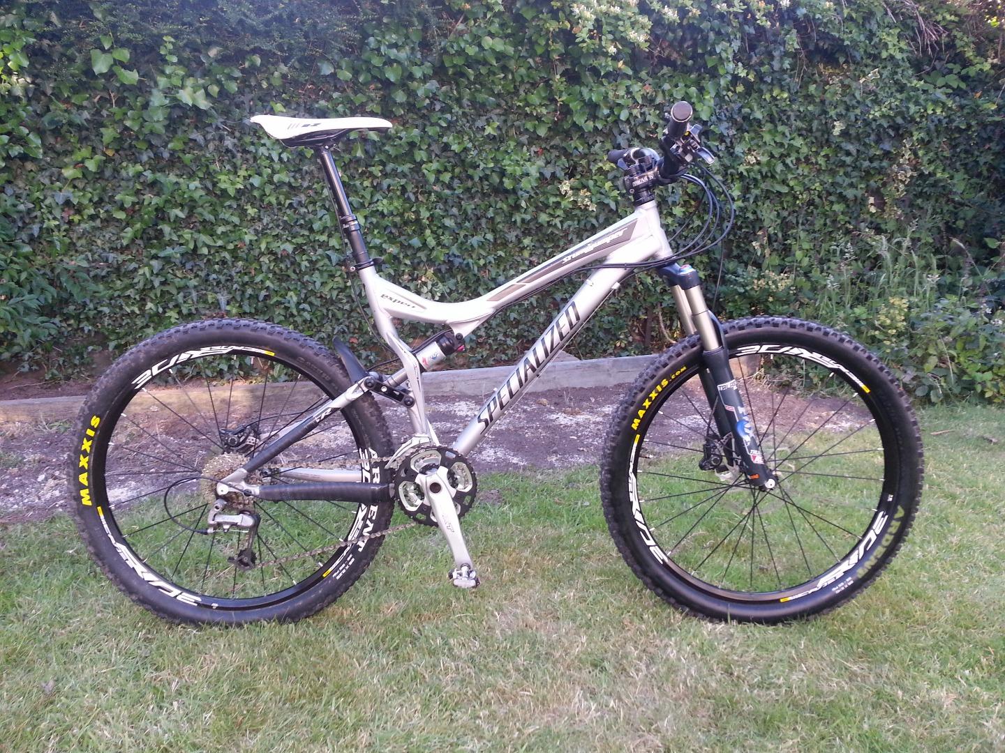 Specialized Stumpjumper FSR: A silver mountain bike with black and yellow wheels, positioned on grass in front of a leafy background. The bike features a front suspension fork, disc brakes, and a sleek design typical of off-road cycling.