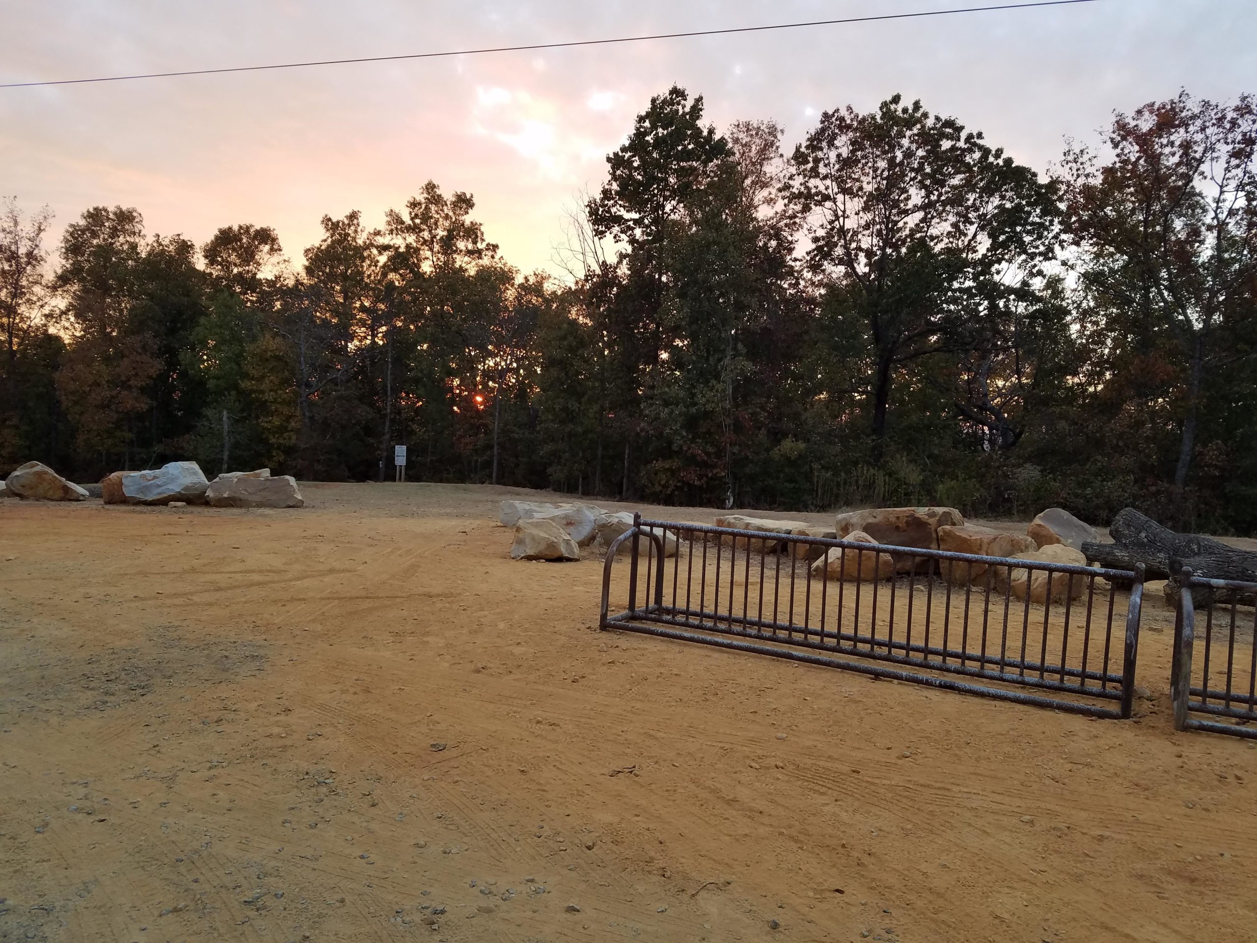 A dirt area bordered by a metallic gate, with large boulders scattered around. In the background, trees display autumn colors, and a colorful sunset can be seen through the foliage. Coldwater Mountain mountain bike trail.
