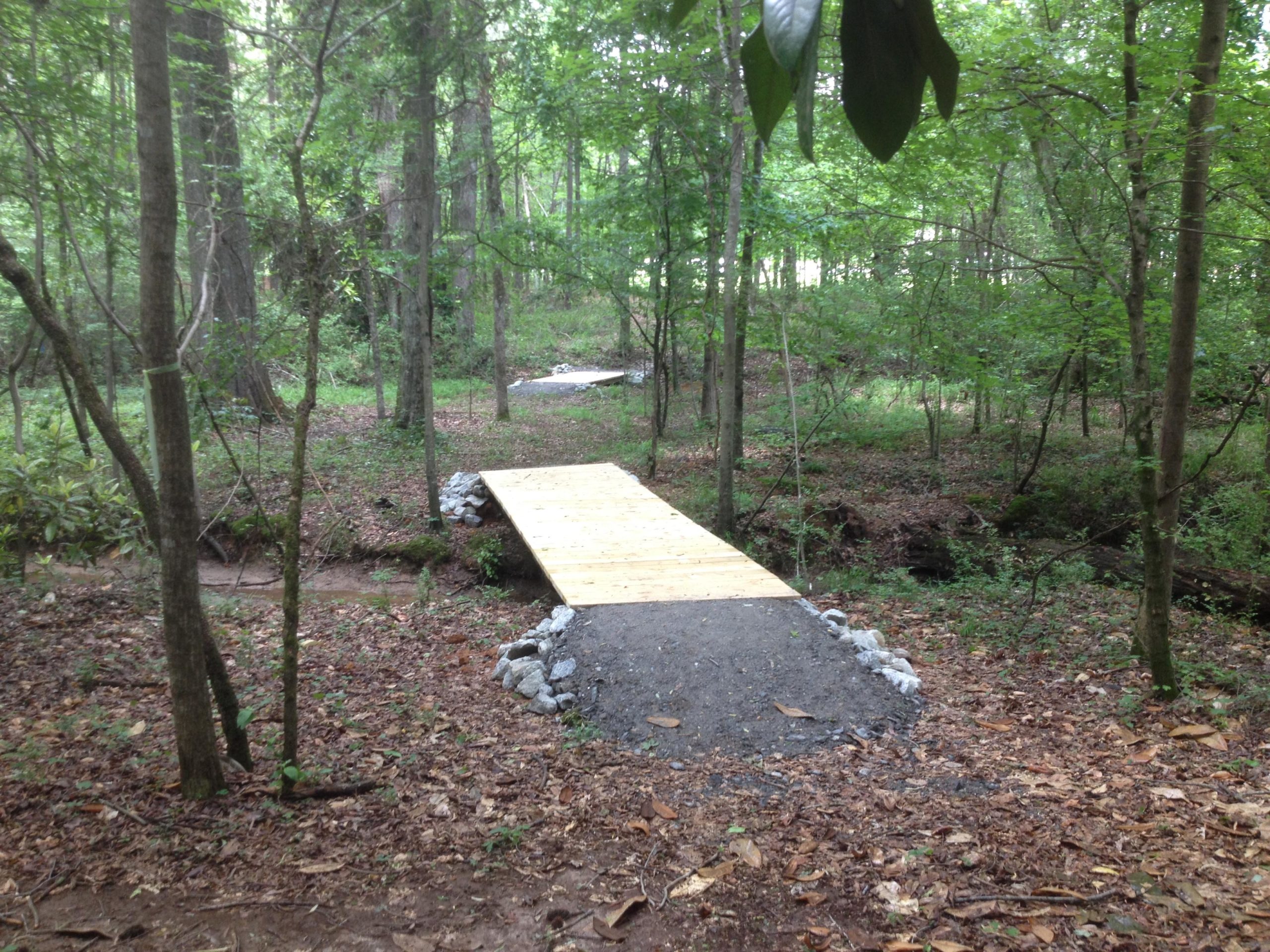 A wooden footbridge spans a small creek in a wooded area, surrounded by lush green trees and underbrush. The ground is covered with fallen leaves, and stones are placed along the edges of the bridge. Austell Threadmill MTB Park (Closed) mountain bike trail.