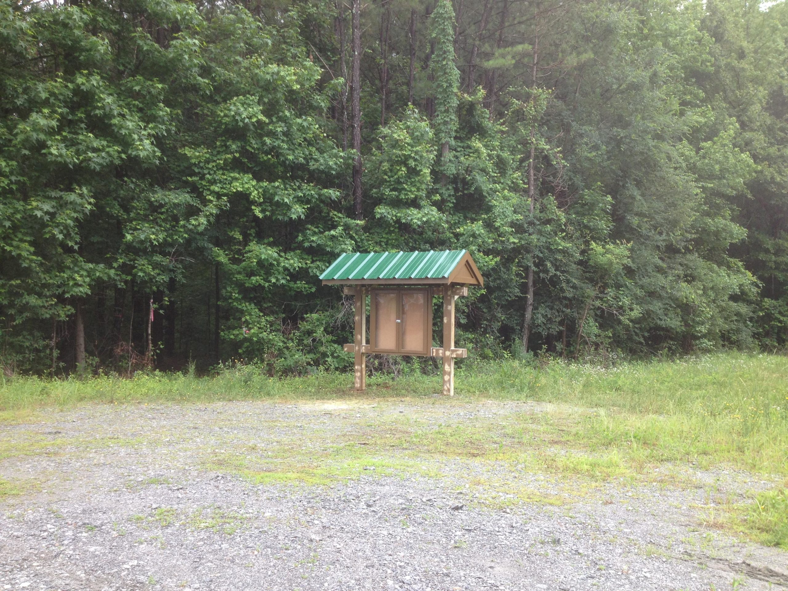 A wooden information kiosk with a green metal roof is located near the edge of a forest, surrounded by gravel and grass. The kiosk stands alone, providing a space for notices or maps, with lush green trees in the background. Austell Threadmill MTB Park (Closed) mountain bike trail.