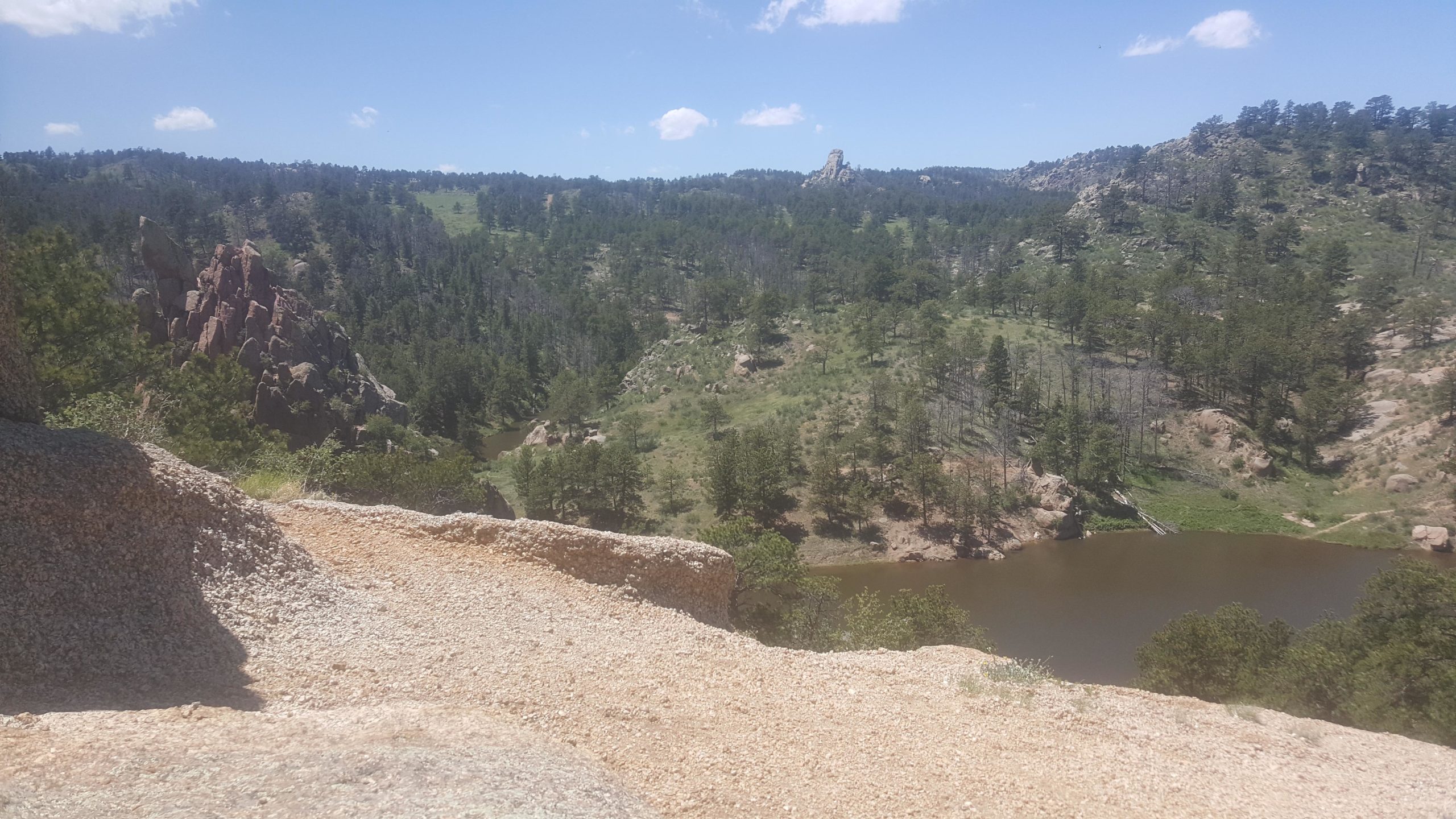 A scenic view of a hilly landscape featuring rocky formations, lush greenery, and a winding river. The sky is bright with a few clouds, creating a picturesque natural setting. Curt Gowdy State Park mountain bike trail.