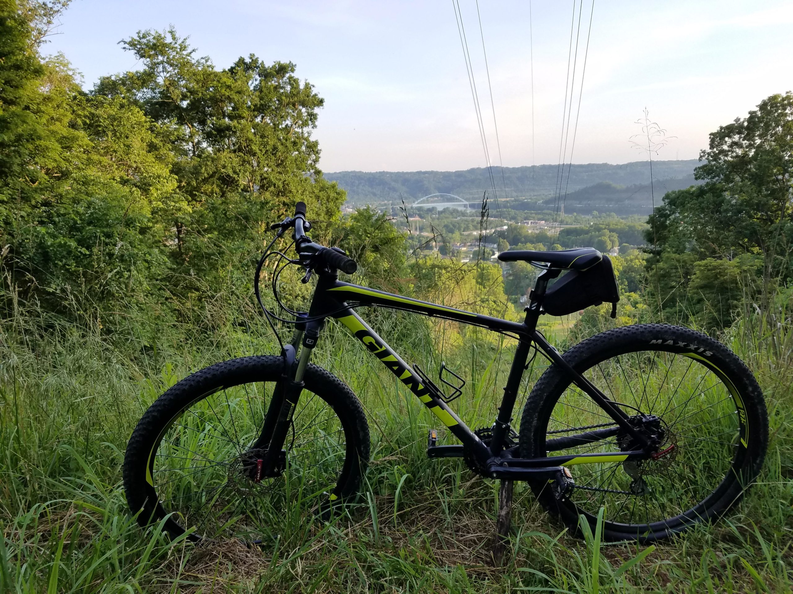 A mountain bike parked on a grassy slope, overlooking a valley with trees and hills in the background. Power lines run across the sky, and the scene is illuminated by soft morning light. Grand Vue Mountain Bike Trail mountain bike trail.