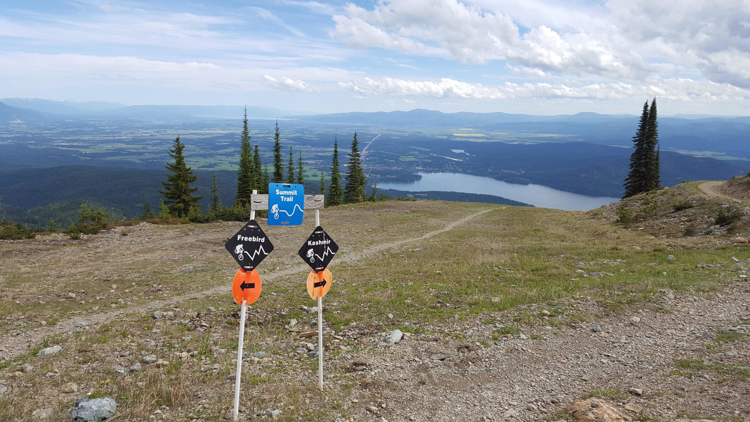 A scenic view from a mountain summit featuring trail signs indicating "Freebird" and "Kashmir" biking trails. In the background, there is a lush green valley and a lake, surrounded by distant mountains under a partly cloudy sky. The foreground shows a gravel path leading into the distance. Whitefish Mountain Resort mountain bike trail.